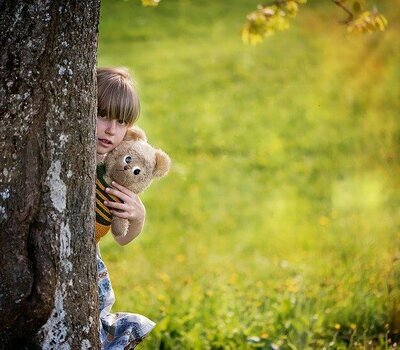 Kind mit Kuscheltier versteckt sich hinter Baum